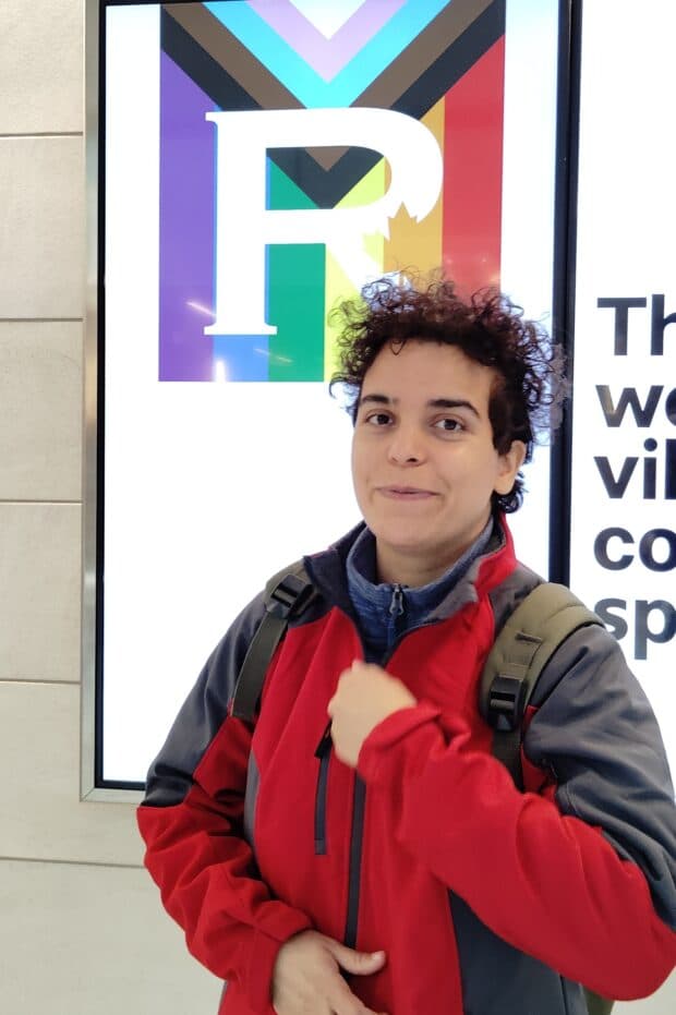 Person standing indoors wearing a red and gray jacket in front of a Pride-themed display.