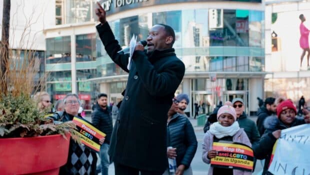Person speaking passionately into a microphone at an outdoor protest, holding a raised fist, with supporters behind holding signs.