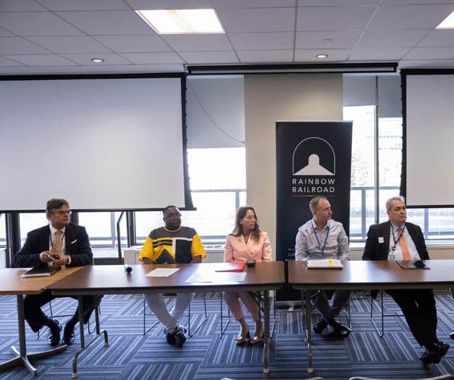 Five panelists seated at a long table with a Rainbow Railroad banner behind them.