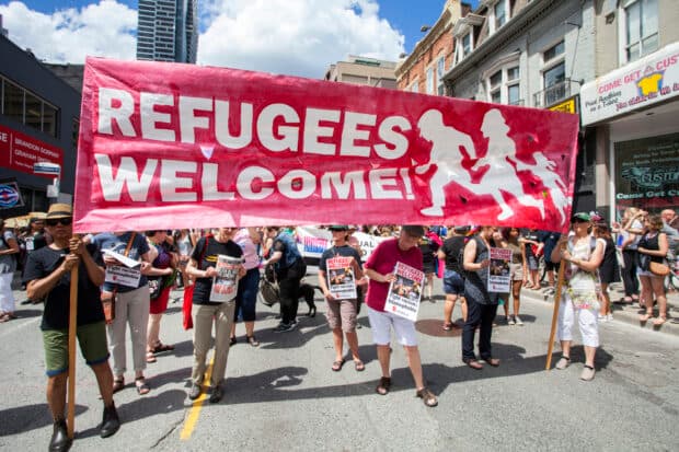 People holds a large red banner reading “REFUGEES WELCOME!” during a street march.