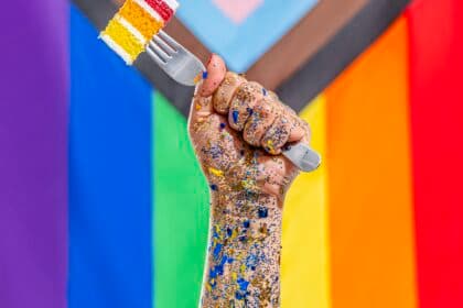 A painted hand holding rainbow cake on a fork in front of a Pride flag.