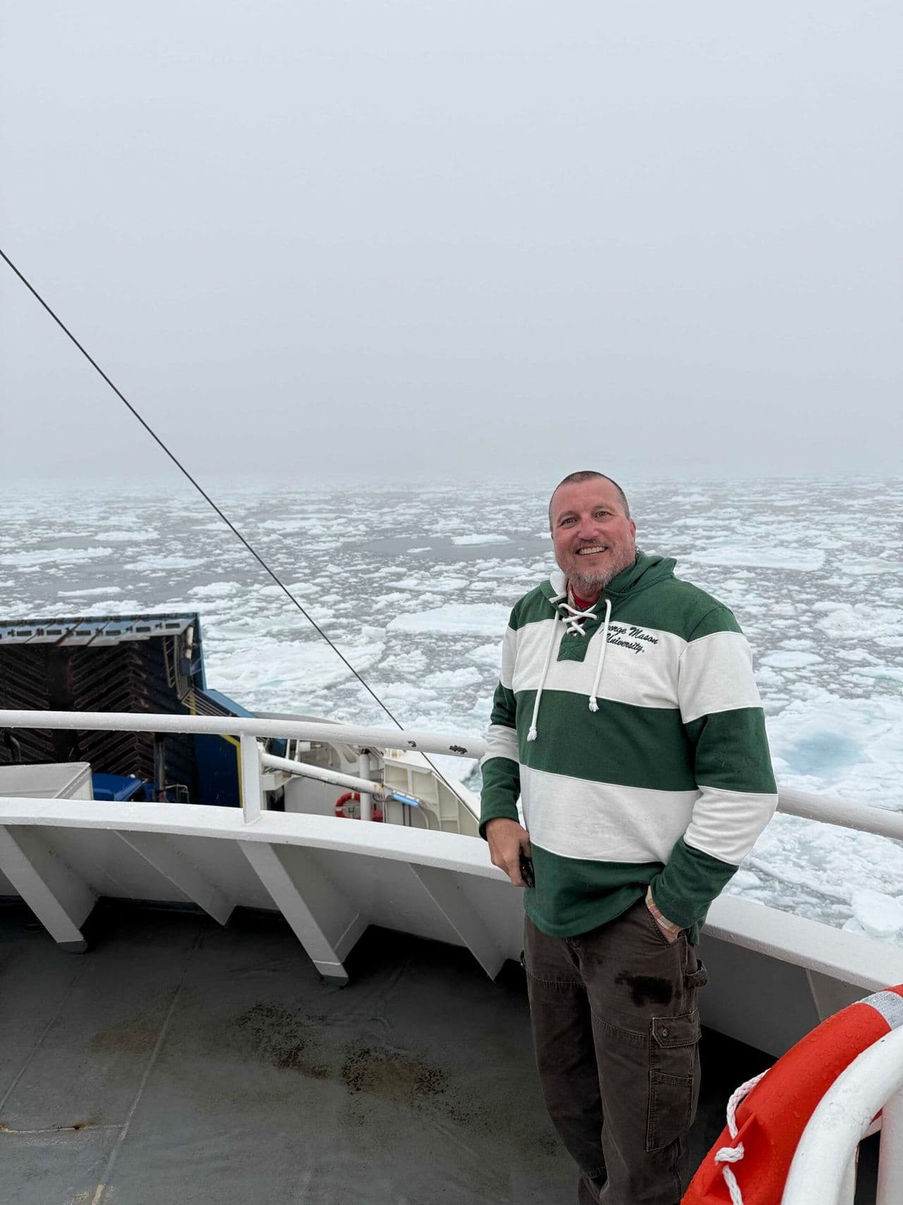 Person standing on a ship deck wearing a green and white striped sweater, smiling with icy ocean waters in the background.