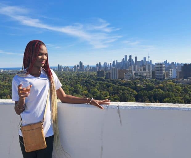 Person with long braids standing on a rooftop holding a drink, with a city skyline in the background.