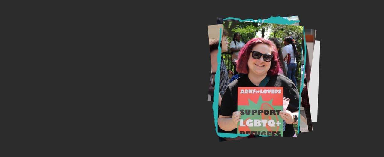 Person with pink hair holding a sign reading ‘Support LGBTQ+ Refugees’ outdoors with people in the background.