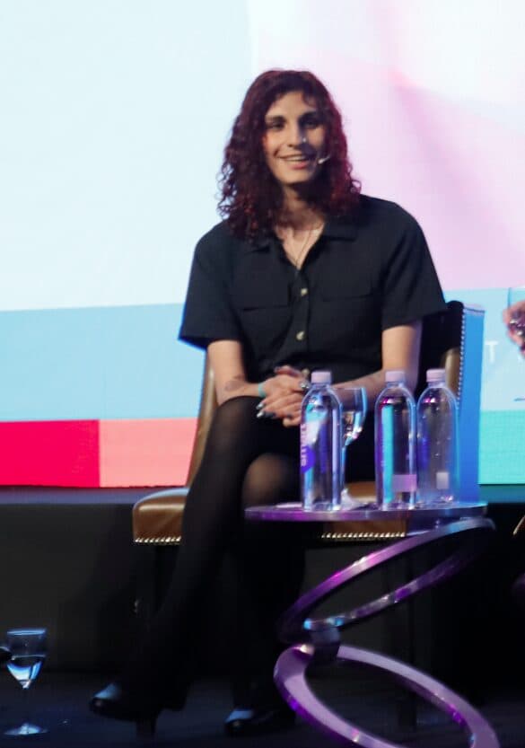 Person with curly hair sitting on a stage panel, wearing a dark outfit and black tights.