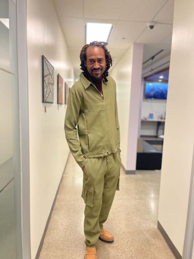A person standing in a hallway wearing an olive green outfit and smiling at the camera.