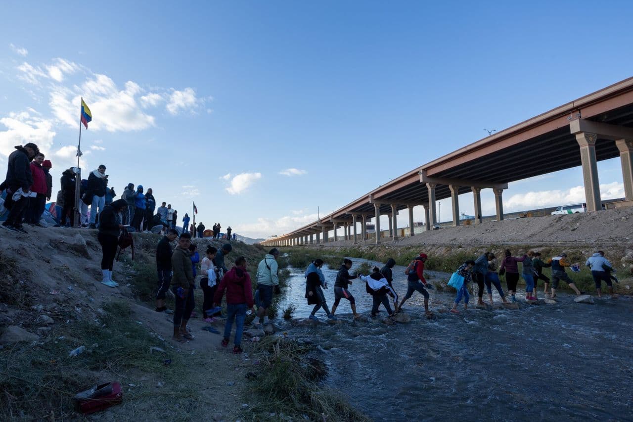 Group of people crossing a shallow river near a large highway bridge.