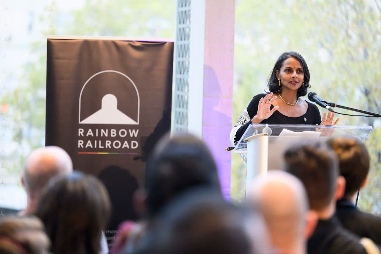 Person speaking at a podium during a Rainbow Railroad event with the organization’s banner beside them.