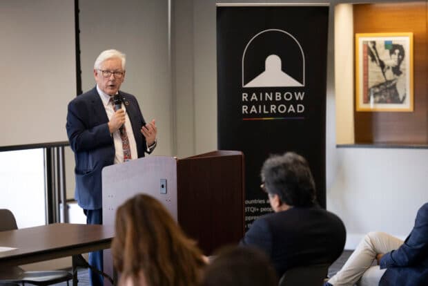 A speaker addresses an audience at a Rainbow Railroad event beside a branded banner.