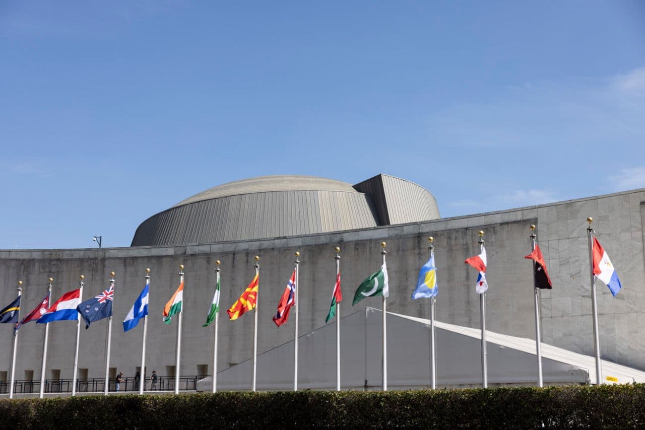 International flags line the front of a large UN building under a blue sky.