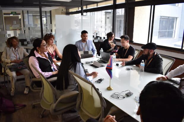 People seated around a conference table in discussion, with a small pride flag displayed at the center.