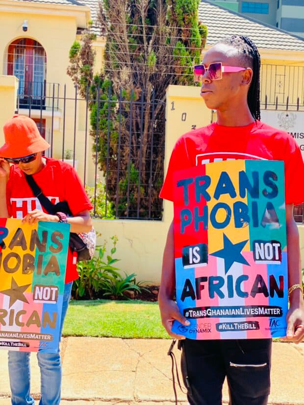 Two protesters in red shirts holding a sign that reads “Transphobia is not African,” outside a government building.