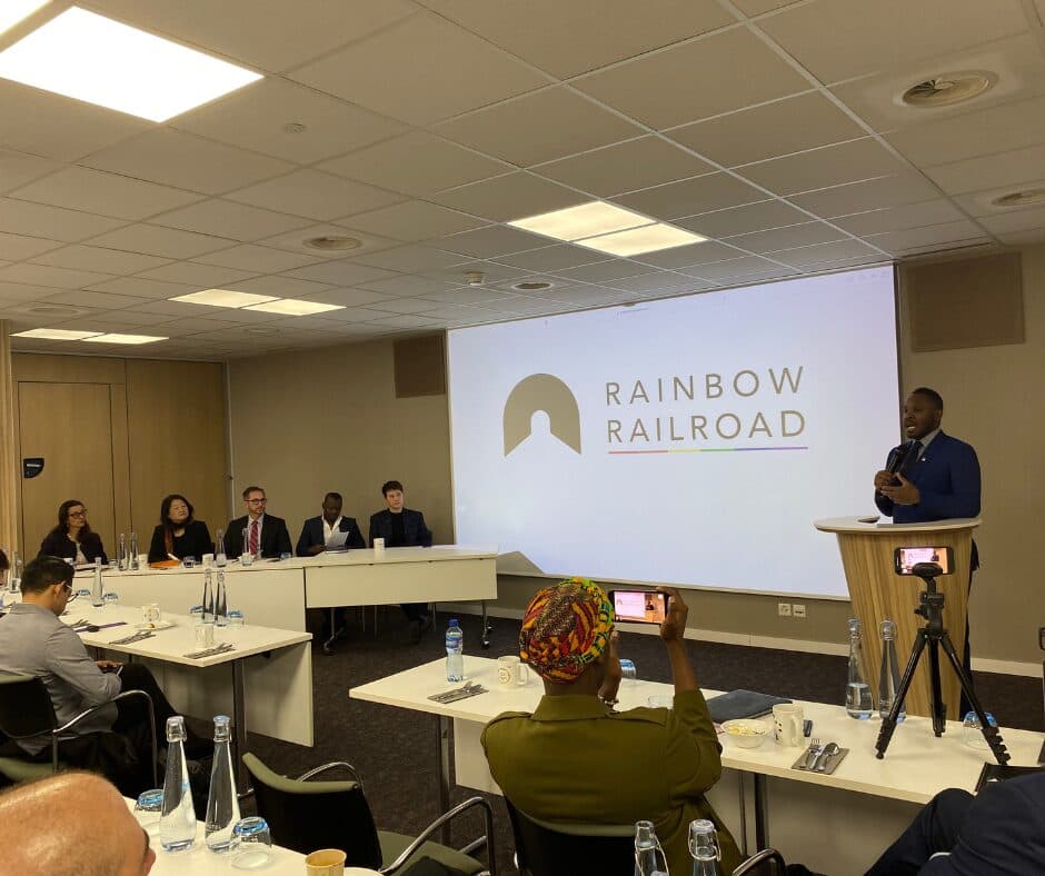 Person speaking in front of a room of people with a screen reading "Rainbow Railroad" in the background.