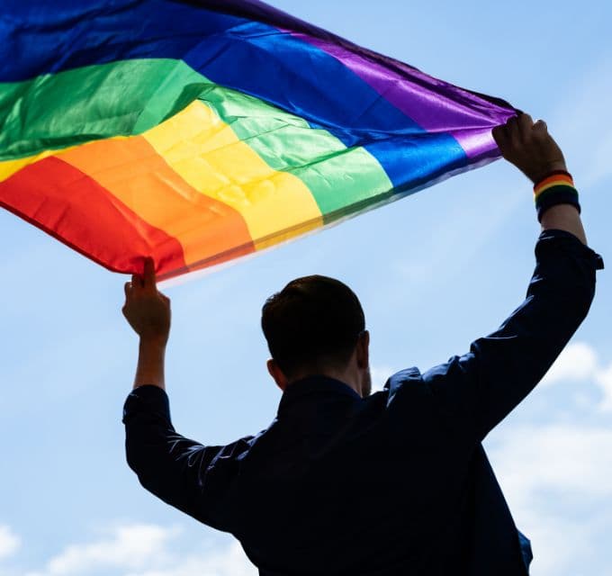 Figure raising a rainbow flag in front of a blue sky.
