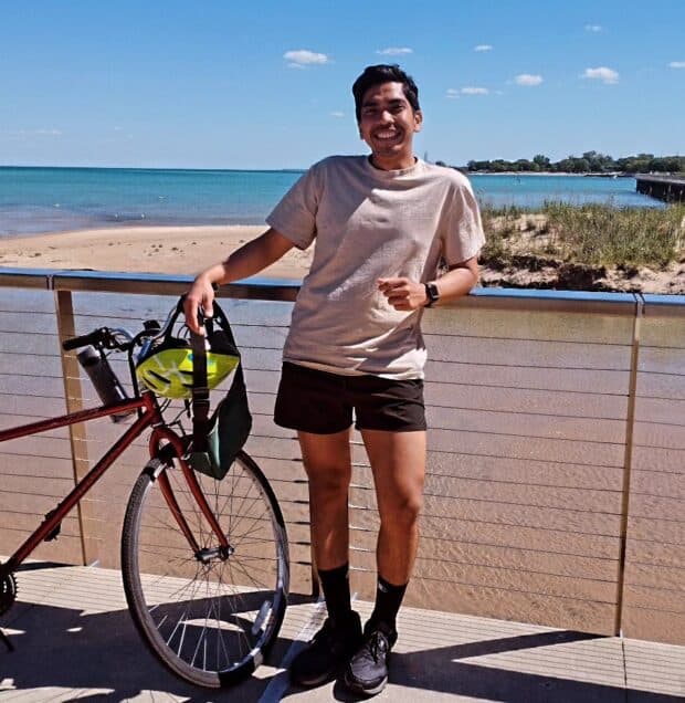 Person standing on a boardwalk beside a red bicycle, smiling with the ocean in the background.