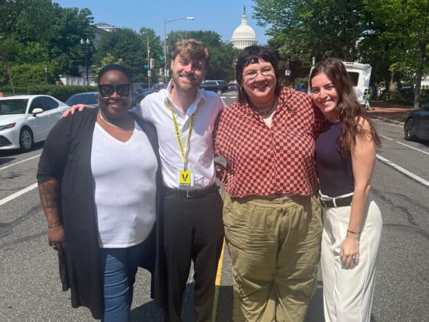 Four people standing together outdoors smiling, with the U.S. Capitol building visible in the background.