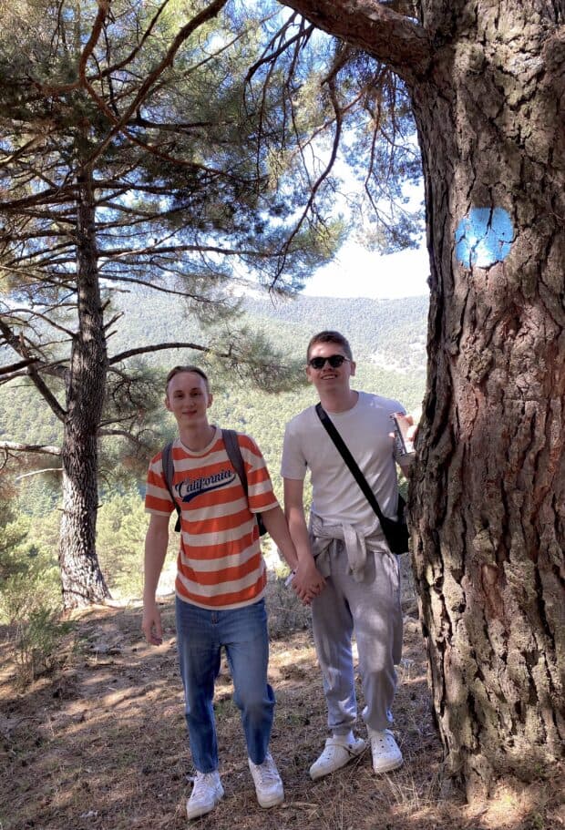 Couple standing outdoors in a forested area, holding hands and smiling near a large tree.