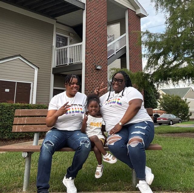 Two adults and a child sitting on a bench wearing matching pride shirts and making peace signs.