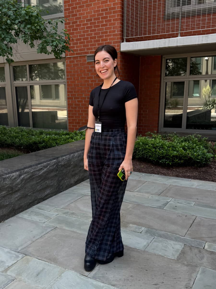 V2 - Person standing outside a brick building smiling, wearing plaid pants and holding a phone.