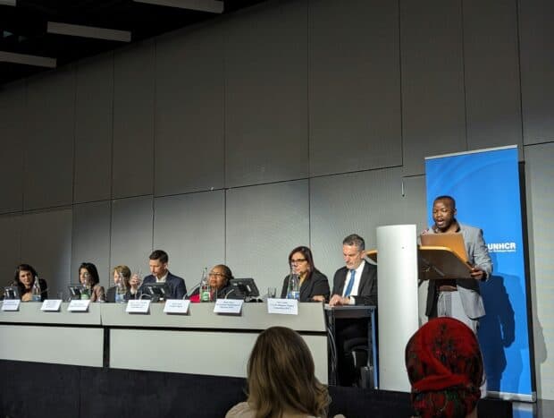 Person speaking on a stage in front of a blue banner with a panel group of speakers beside them.