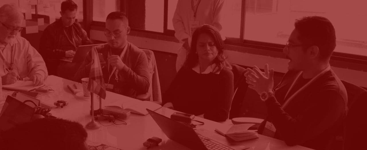 People seated at a table in discussion, with notebooks and laptops in front of them, under a red color overlay.