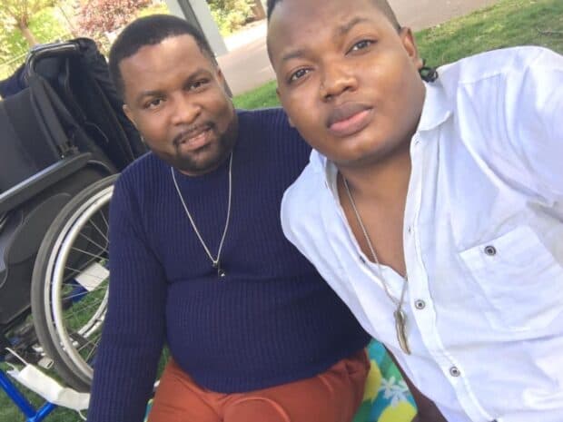 Two people sitting on a blanket outdoors beside a wheelchair, smiling at the camera.