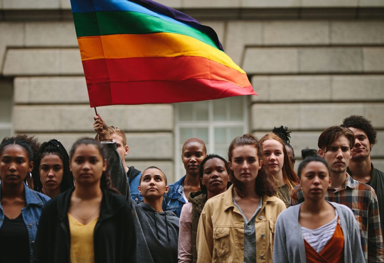 A group of people stand together as one person raises a rainbow Pride flag above them.