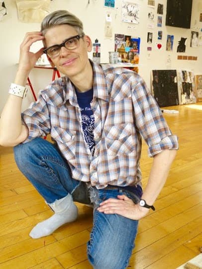 Person kneeling on a studio floor, smiling and wearing glasses and a plaid shirt.